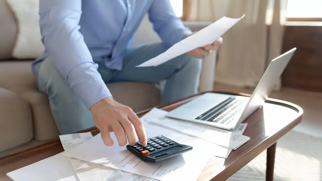 pessoa de camisa azul sentada em frente a uma mesa com um notebook, mexendo em uma calculadora para serviços contábeis